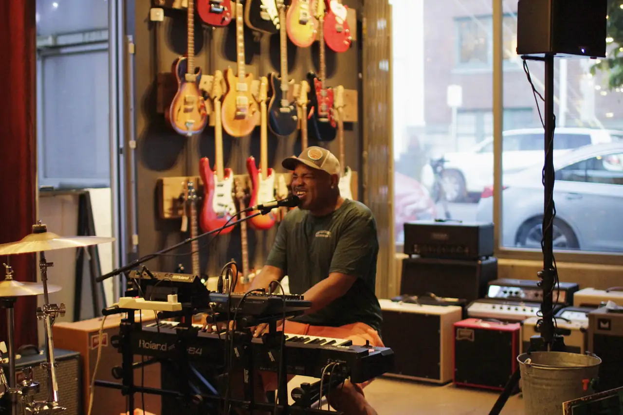 Singing at the keys in a guitar shop, amps stacked by the front window