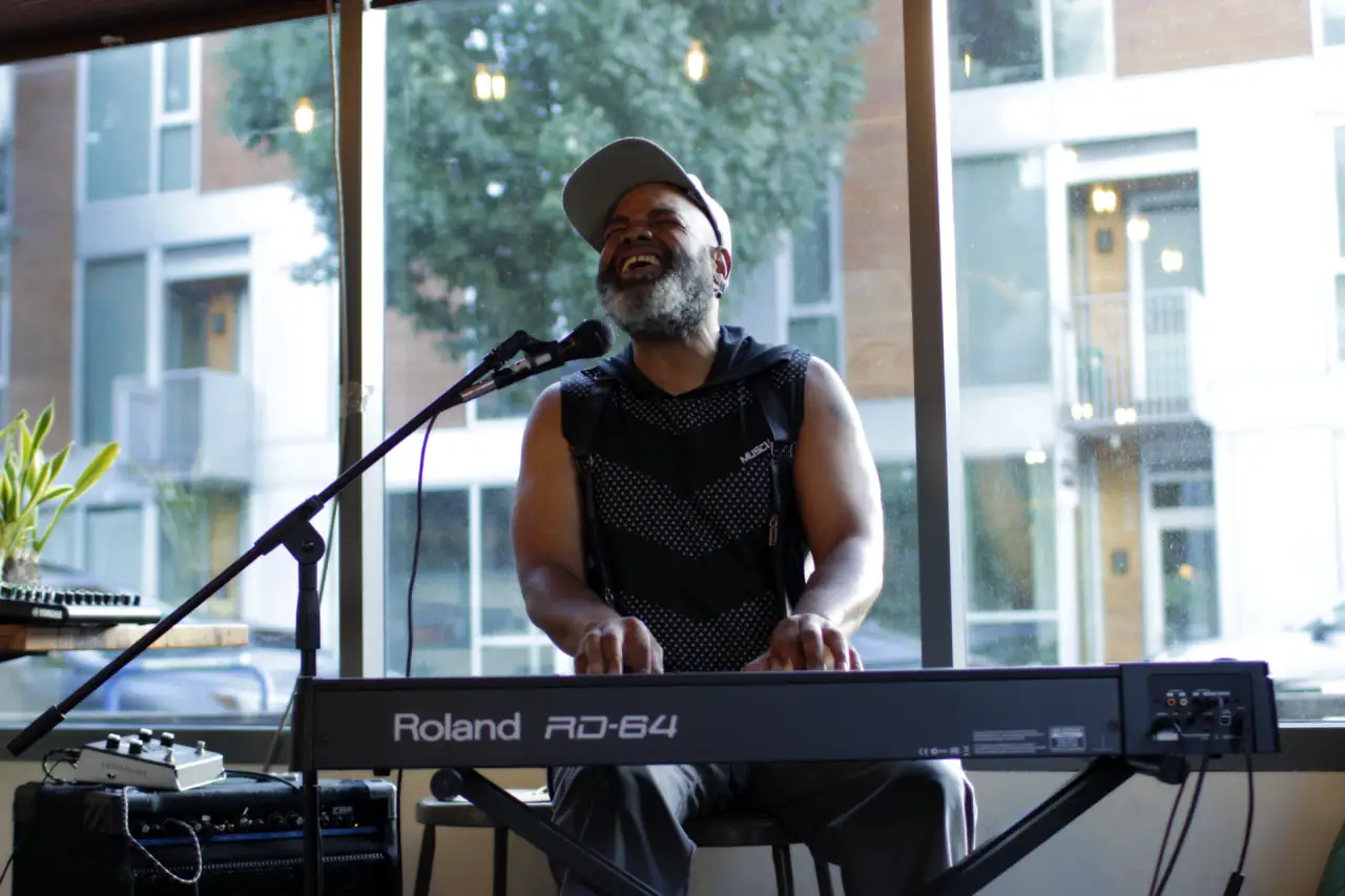 A pianist singing at a Roland keyboard by a sunlit window