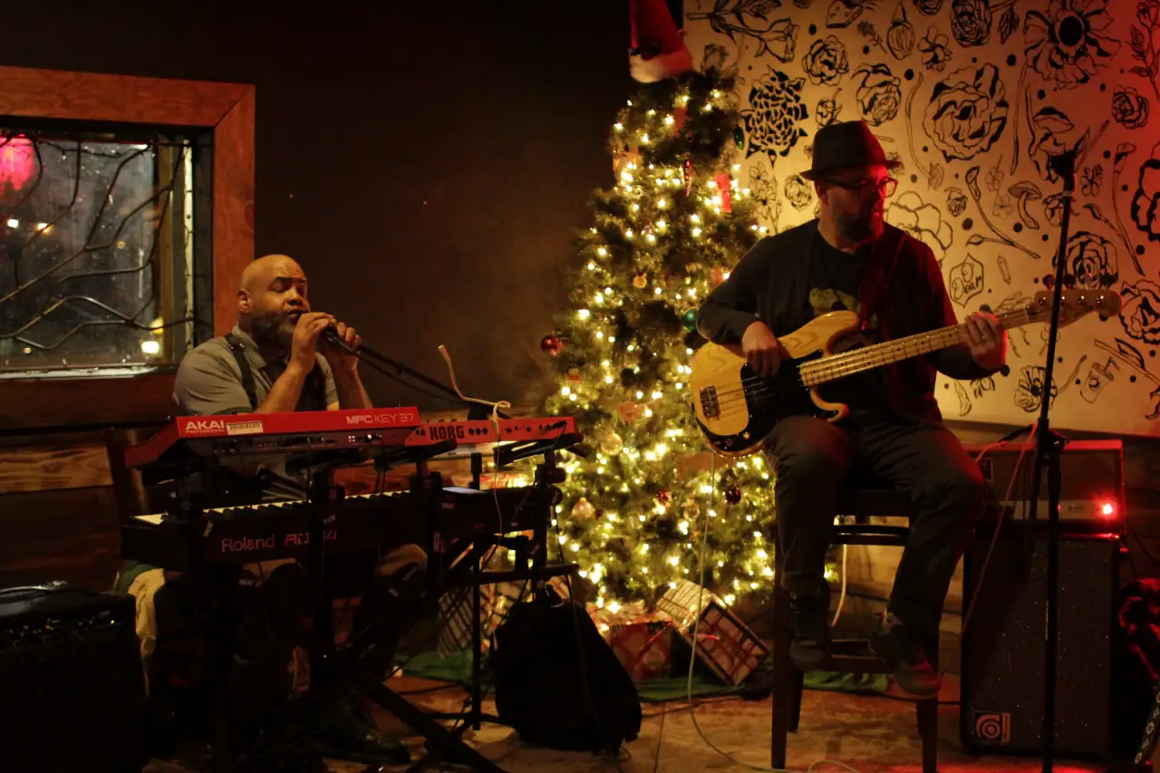 Keys and bass playing a holiday show beside a lit Christmas tree